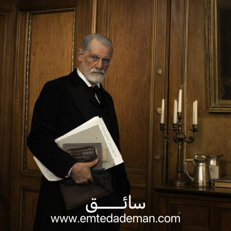 مردی سالخورده با ظاهری رسمی که پوشهها و نوشتههایی را در دست دارد و در فضایی کلاسیک و چوبی ایستاده است؛ تصویری نمادین از ذهن تحلیلگر، اقتدار فکری و ریشههای روانکاوی.
